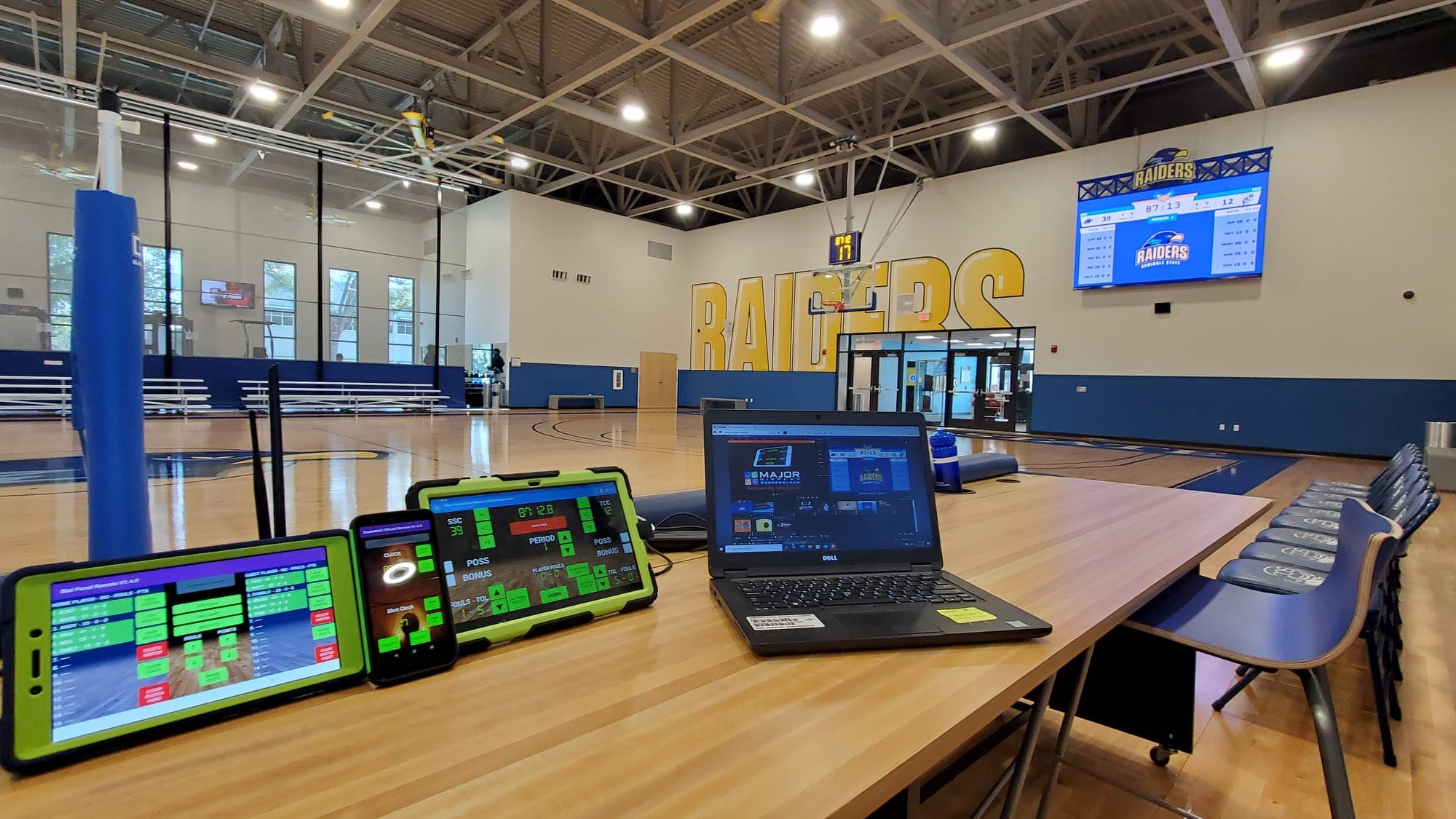 Several SCB3000 controllers, a phone, and a laptop on one scorer's table, with the facility's video boards on the wall